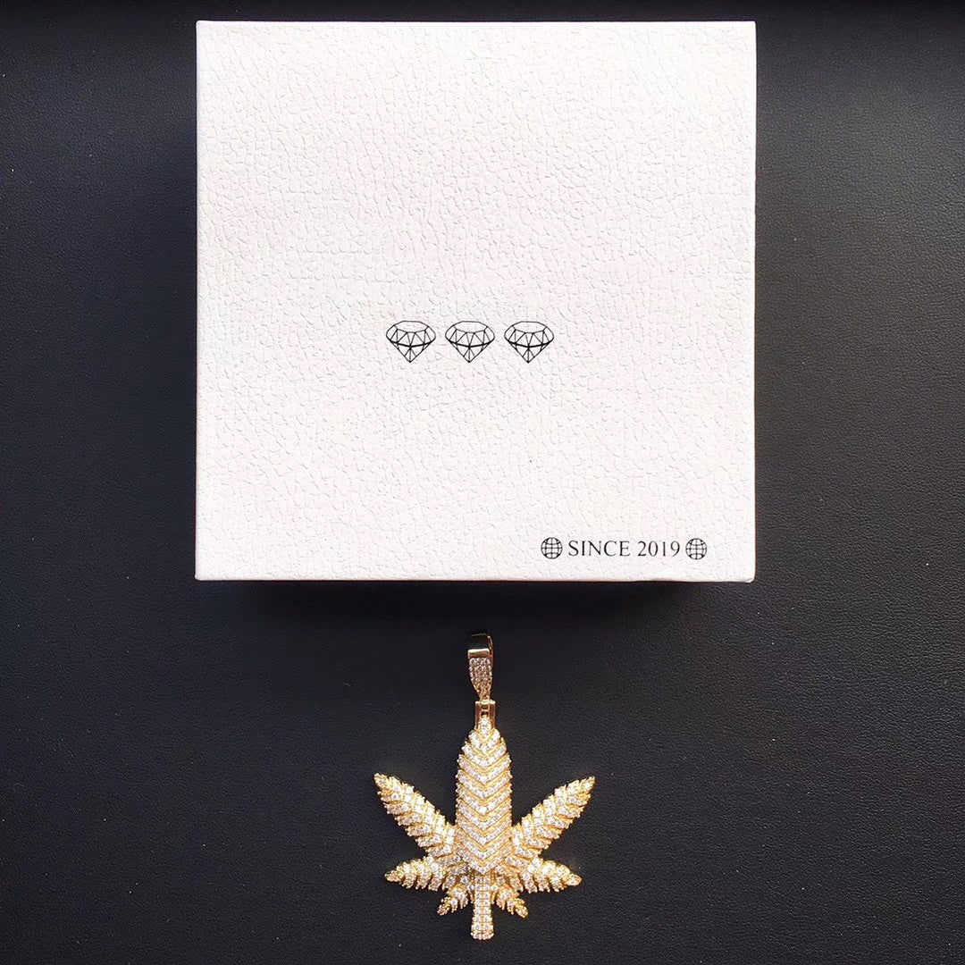 DatNewDrip iced out pot leaf pendant gold plated with clear stones displayed below white gift box on black background